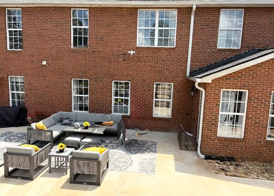 A spacious concrete back patio featuring a modern grey sectional sofa set with yellow accent pillows against a red brick house.