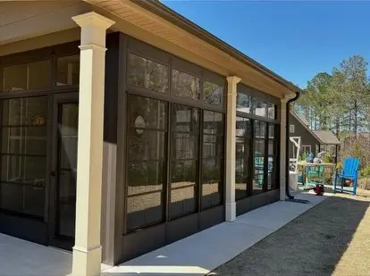 A modern bronze Sunspace sunroom enclosure installed on a concrete patio with cream pillars.