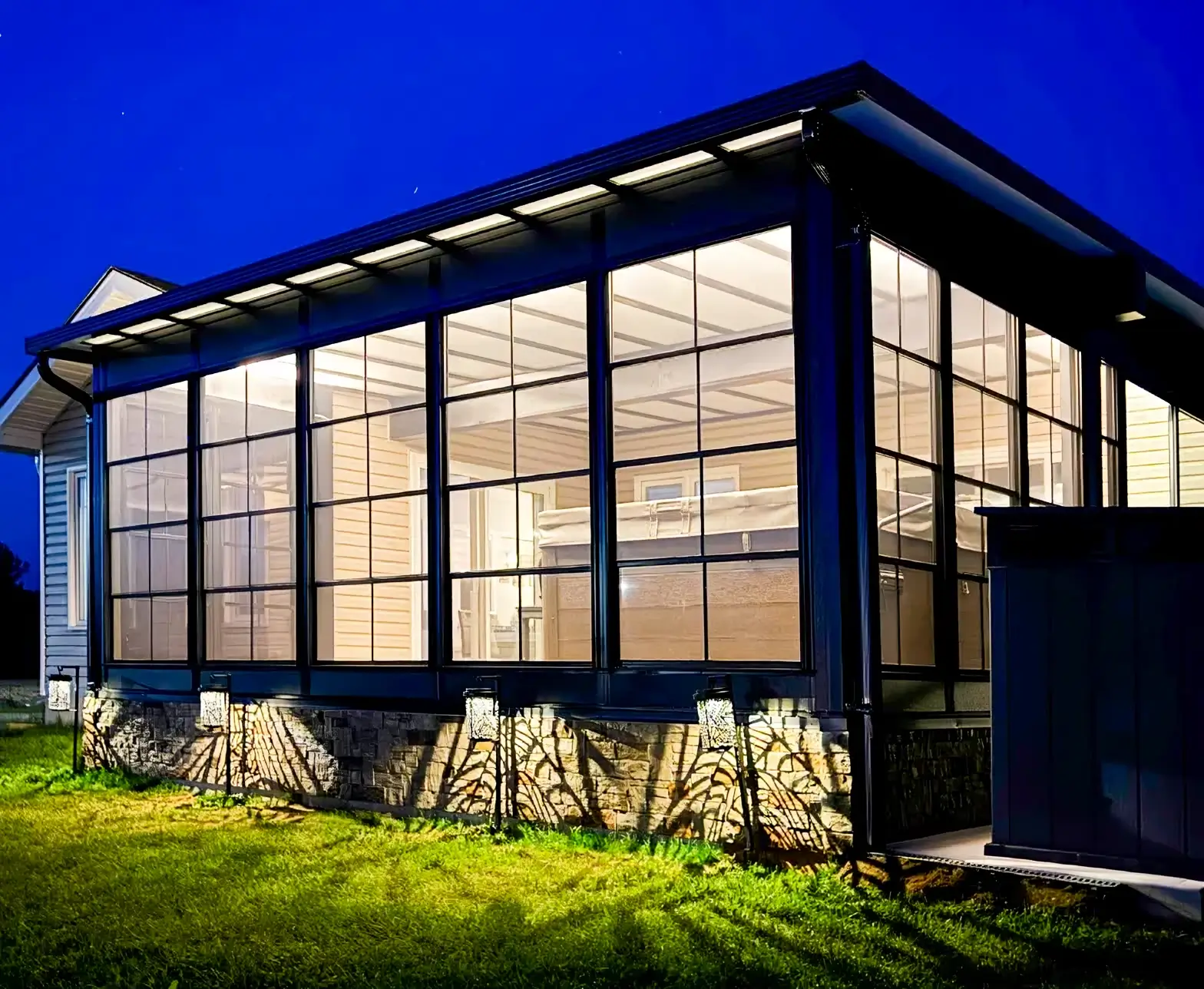 An illuminated evening view of a Sunspace Sunrooms 200 enclosure featuring dark frames and a hot tub