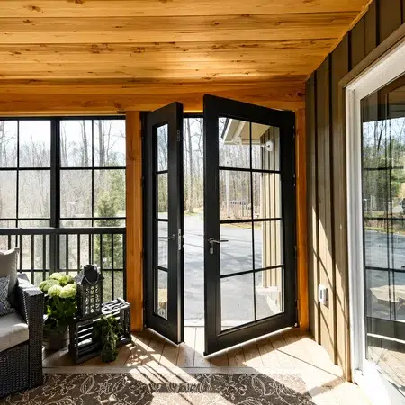 Inside a sunroom with a wood ceiling, looking out through open black double French doors onto a driveway and wooded yard.